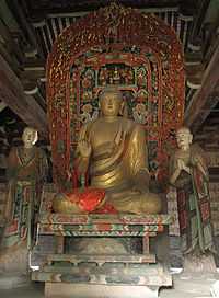 Shown inside the high-ceilinged hall is  a large, gold-colored statue seated cross-legged  on an ornately decorated throne, flanked by two figures in richly coloured robes.