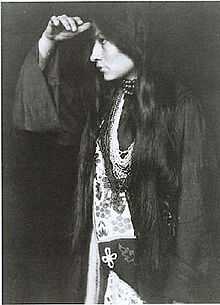 Photo of Zitkala Sa in profile, wearing Native American dress, with long dark hair hanging below waist, holding hand at forehead and looking into the distance