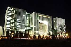 A tall modern office building of three flat-roofed rectilinear blocks joined by smaller hyphens. It is seen at night, with some of its windows lit.