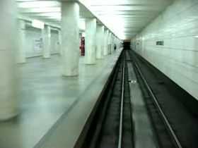 A silent video shot from the perspective of the driver's cab of a metro train. It travels in a dark tunnel and then stops at the underground station, which is lit up in white and supported by the pillar-trispan structure.