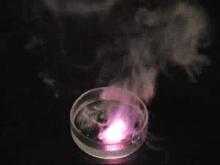 A piece of potassium metal is dropped into a clear container containing water and slides around, burning with a bright pinkish or lilac flame for a short time until finishing with a pop and splash.