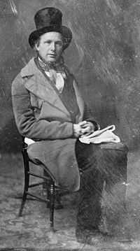 A black and white photograph, slightly damaged, showing a seated man wearing an overcoat and top hat