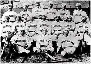 Baseball players are posing for a photograph, six men standing, seven men sitting on chairs, and three are sitting on the ground.