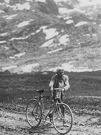 A man walking, holding, to his bicycle, on a sandy road.