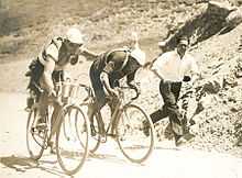 Two cyclists on bicycles, one cyclist with his hand on the back of the other cyclist. In the background, one man is running in the same direction.