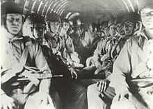 Men with helmets sit in an aircraft with weapons held across their chests, strapped into parachutes