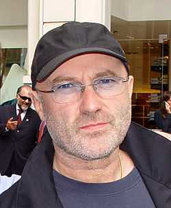 A picture of a man with glasses. He wears a black cap and a black coat over a dark blue shirt.