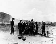 A large artillery gun fires as several soldiers look on