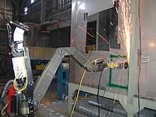 A robot with a metal arm is using its grinding wheel to cut into a wall, sending a shower of sparks upward.