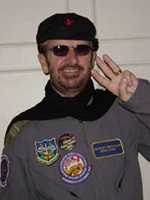 A colour photo of Starr, who is holding two fingers up in the style of a "peace sign". He is a wearing a dark beret, sunglasses and a grey windbreaker with several patches on the front.