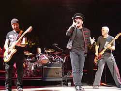 Four men performing on a stage in front of a crowd; two are standing at the front of the stage holding guitars, one in the center is holding a microphone, and one is sitting behind a drum set. Audio equipment and microphone stands can also be seen on the stage.