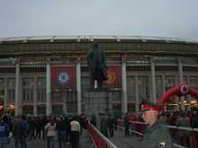 A wide shot of a large, bronze statue of Lenin on a tall plinth. In the background is a stadium draped with red banners, one with the Manchester United crest and one with the Chelsea crest. People are walking towards the stadium and a Russian policeman is in the foreground.