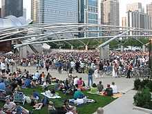 A large number of people are seated on the grass facing a bandshell with an even larger crowd across a wide sidewalk. The bandshell is framed by shiny curved metal and a trellis is over the larger crowd. Skyscrapers are in the background.