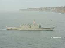 A large grey warship underway. Flags are being flown from the ship's mast, and white-uniformed people line the decks. Brown cliffs line the horizon for the right two-thirds of the image