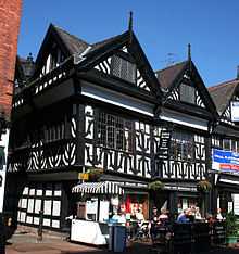 A black-and-white building in a corner position, with two gables to the front and one visible to the left side. On the street in front is a pavement café.
