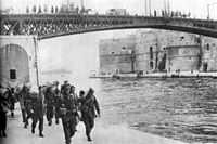 Soldiers carrying kit-bags approaching the photographer, while walking on an embankment next to a river with steel girder bridge in the background