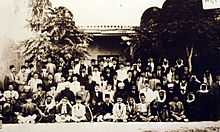 Large group of men in front of building and trees