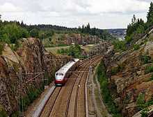 A four-car multiple unit running along a double-track line between two cliffs; in the background is a motorway.