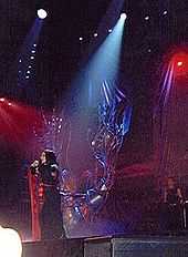 Image of a brunette woman in a black-and-red kimono moving to the right of a stage with red-lit backgrounds and a metallic tree like structure in the middle. A single source of light falls on her.
