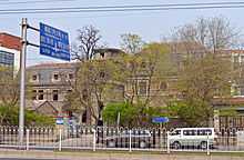 The building in the infobox and another, similar one to its right, seen from across a street in front divided by a white metal fence, with a blue road sign in Chinese and English at left. The gate of the fence is open and trees, with leaves slowly opening, are behind it.