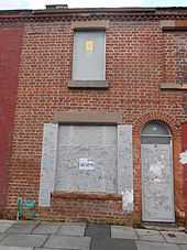 A colour photograph of a shabby looking, red bricked house with boarded up windows and doors