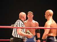 Three adult males standing in a wrestling ring. One is bald wearing a black and white striped shirt holding a championship belt. Another is staring at him wearing blue wrestling gear. The third is doing the same but is wearing black wrestling gear and is bald.