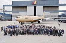 Crowd of people assembled in front of unpainted aircraft. A tall building serves as the backdrop for the photograph