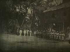 A line of soldiers in tropical uniforms stand in front of a hut in a jungle clearing. In front of them stand a group of five men facing towards the camera