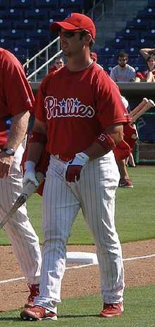 A man wearing a red baseball jersey and cap and white pinstriped baseball pants walking on a grass field