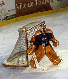 An ice hockey player standing partially crouched in goals. He is wearing a helmet, gloves and leg pads and a black and orange uniform.