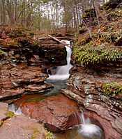 A narrow falls resembling a horsetail is in a chute carved in layers of rock, which then flows into a large pool at the base. The surrounding rock has green bushes and a brown picket fence is visible above the falls.