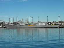 Adelaide tied up alongside at a wharf. The majority of her communications and radar masts have been removed, she carries no weapons, and several large squares have been cut into the ship's hull.