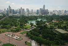 A park with many trees and a lake; a bronze standing statue in front of the park; many buildings in the background