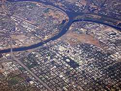 High angled aerial shot of a developed city; a suburban grid dominates the lower half of the image. A river bisects the city from the left before forking; the first fork continues up and to the right edge of the image; the second curves up and around to finish on the left, enclosing industrial units and other domestic properties.