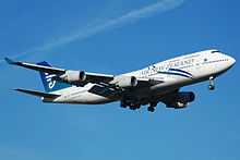  An Air New Zealand 747-400 with its landing gear down and flaps down.  The aircraft is mostly white and blue aircraft and in-flight against a blue sky. On each of the two wings are two engines.