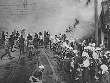 Black and white photo of women standing on a street passing buckets along a chain of people towards a building on fire. Other people are climbing a ladder from the street into the building.