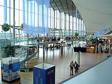 Inside a terminal, with a multi-story glass wall to the left; the open space is filled with signs, chairs, small store and amenities