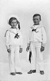 A black and white portrait of two young boys. They are standing alongside one another and are dressed in whites sailor uniforms. The shorter boy on the left is smiling and wearing a cap, while the boy on the right has a blank expression and is holding a cap in his left hand.