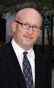 Photo of a balded Caucasian male wearing glasses, a black coat over a white collared shirt, and a blue tie with white and maroon diamonds.