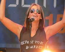  A young woman with blonde hair singing into a microphone on a stand while wearing a black top with the wording 'Prepare to be boarded' and dark sunglasses.