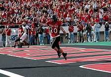 Texas Tech Red Raider football player stands with the a football in the endzone in the foreground with fans in the stands in the background cheering.