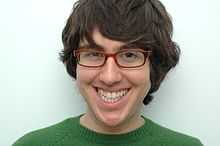 Amir Blumenfeld with long hair, red glasses and a green sweater, grinning before a white background