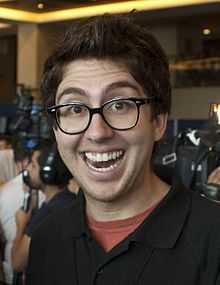 Amir Blumenfeld smiling widely, in front of a crowd of journalists with cameras, wearing a black polo shirt and horn-rimmed glasses