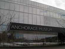 The mirrored facade of a building is seen with a stainless steel nameplate reading Anchorage Museum.