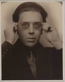A sepia photograph of a man dressed in a suit. He is wearing a pair of semi-transparent sunglasses and has both hands raised to either side of his head. The photograph is surrounded by a white border.