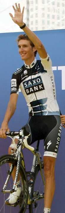 A cyclist in a black and white jersey with blue trim sits atop his bicycle, waving with his left hand and smiling.