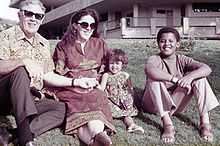 A young boy (preteen), a younger girl (toddler), a woman (about age thirty) and a man (in his mid-fifties) sit on a lawn wearing contemporary c.-1970 attire. The adults wear sunglasses and the boy wears sandals.