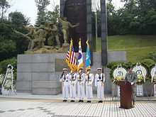A line of sailors in white dress naval uniforms in front of a large monument while another soldier in green stands at a podium beside