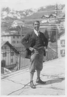 A black and white photo of a man standing on a street. He has his hands on his hips and is smiling towards the camera.