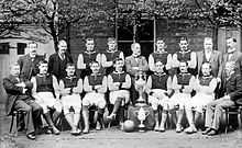 A black and white photograph showing two rows of people in front of a building; the front row are seated with two trophies in the middle, the rear row standing.